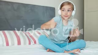 Child girl sitting on the bed and listening music in headphones from smartphone cell