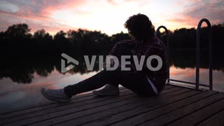 Depressed young man sitting and pondering about life with sunset lake view